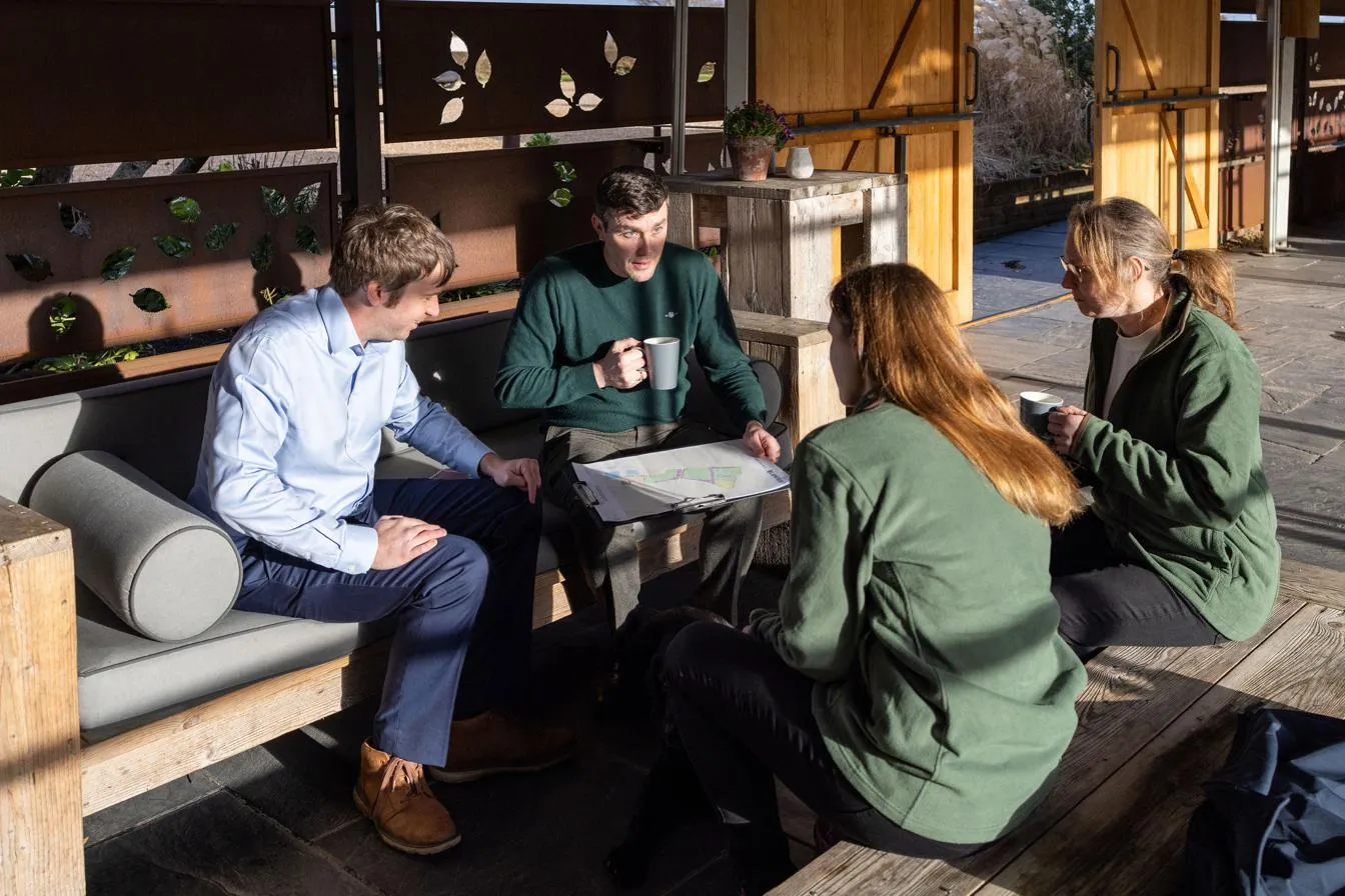Four people sitting outdoors with mugs of tea, discussing a habitat plan spread on a clipboard.